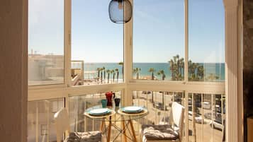 A balcony with a view of the beach, a glass table, two chairs, and a hanging light fixture.