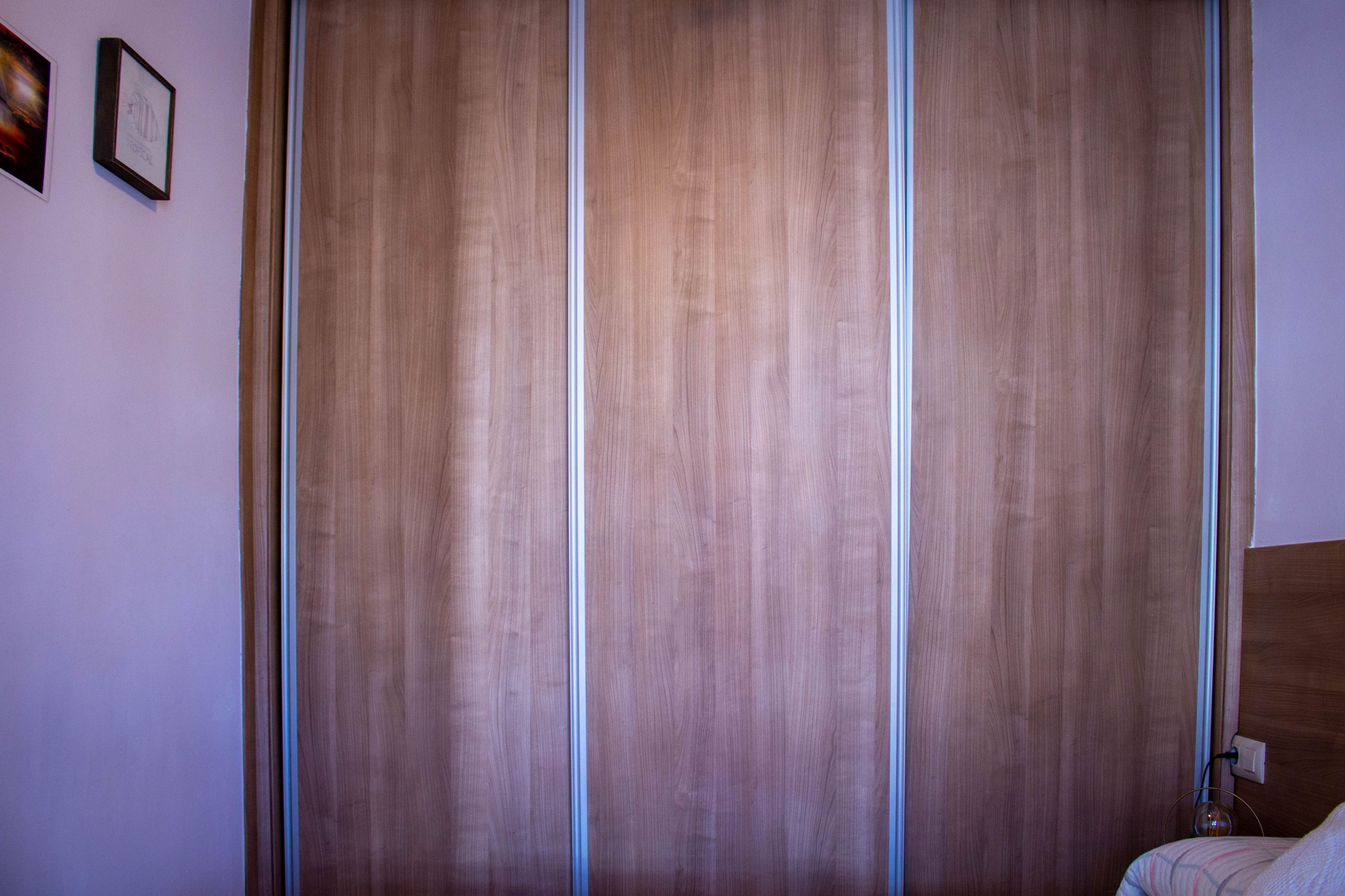 A room with a wooden wardrobe, a bed with a headboard, and a wall-mounted light fixture.