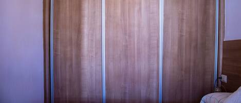 A room with a wooden wardrobe, a bed with a headboard, and a wall-mounted light fixture.