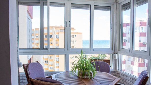 Une pièce avec une table et des chaises en bois, une plante en pot et de grandes fenêtres donnant sur un immeuble et la mer.