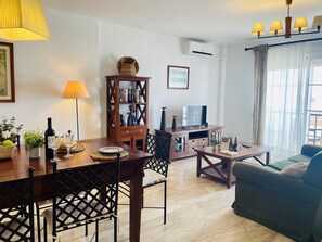 A living room with a wooden dining table, chairs, a sofa, a TV stand, and a bookshelf. - Albatros 3 by At Home Costa del Sol (Algarrobo)