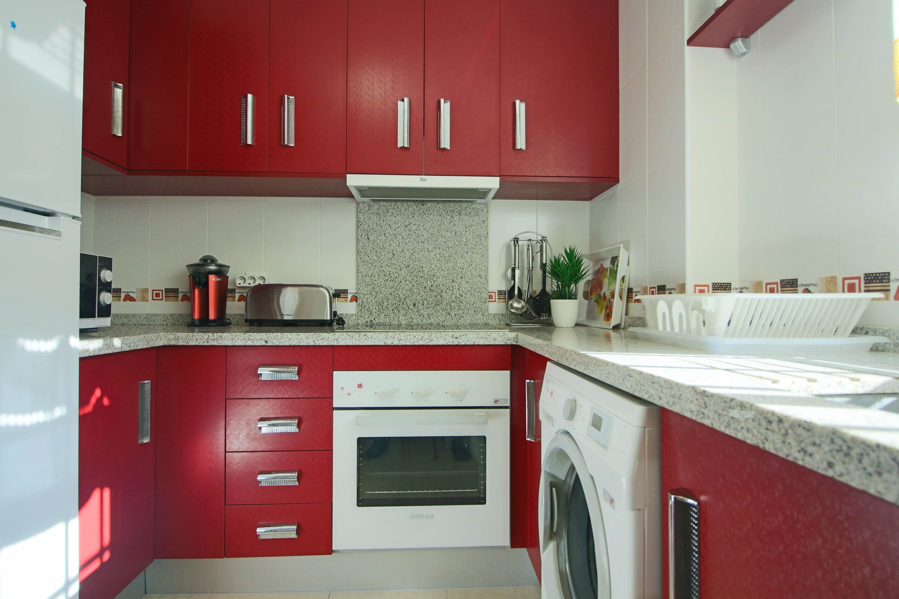 A kitchen with red cabinets, a built-in oven, a washing machine, and a countertop with various appliances.