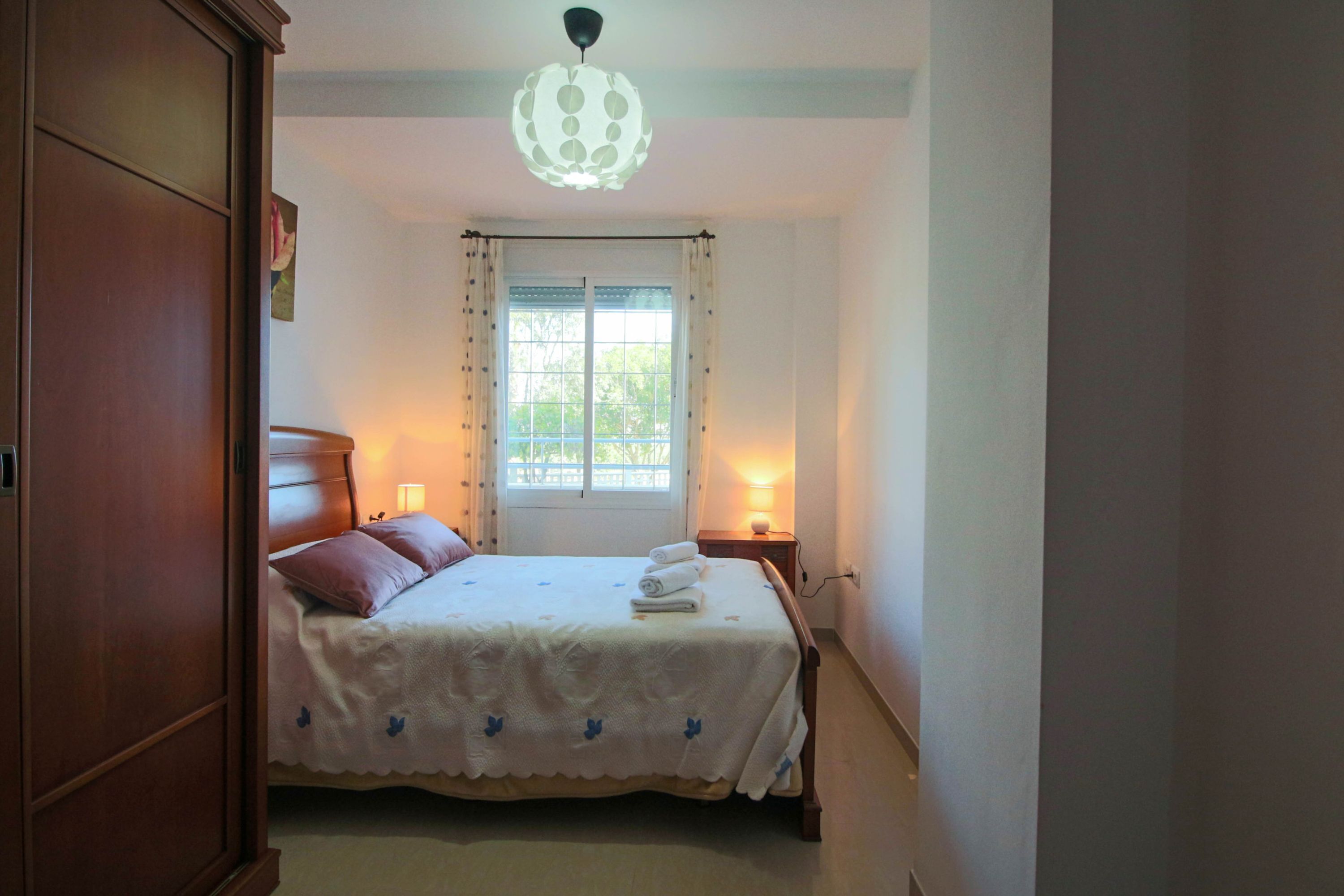 A bedroom with a bed, bedside lamps, a window with curtains, and a wooden wardrobe.