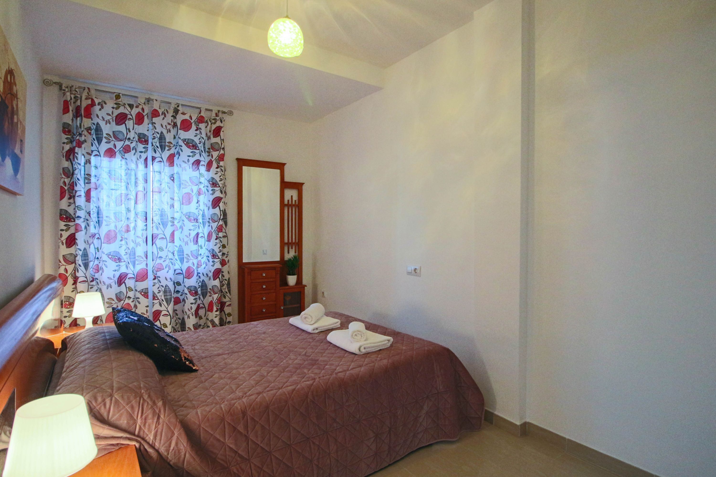 Un dormitorio con cama, mesita de noche, lámpara, espejo y ventana con cortinas.