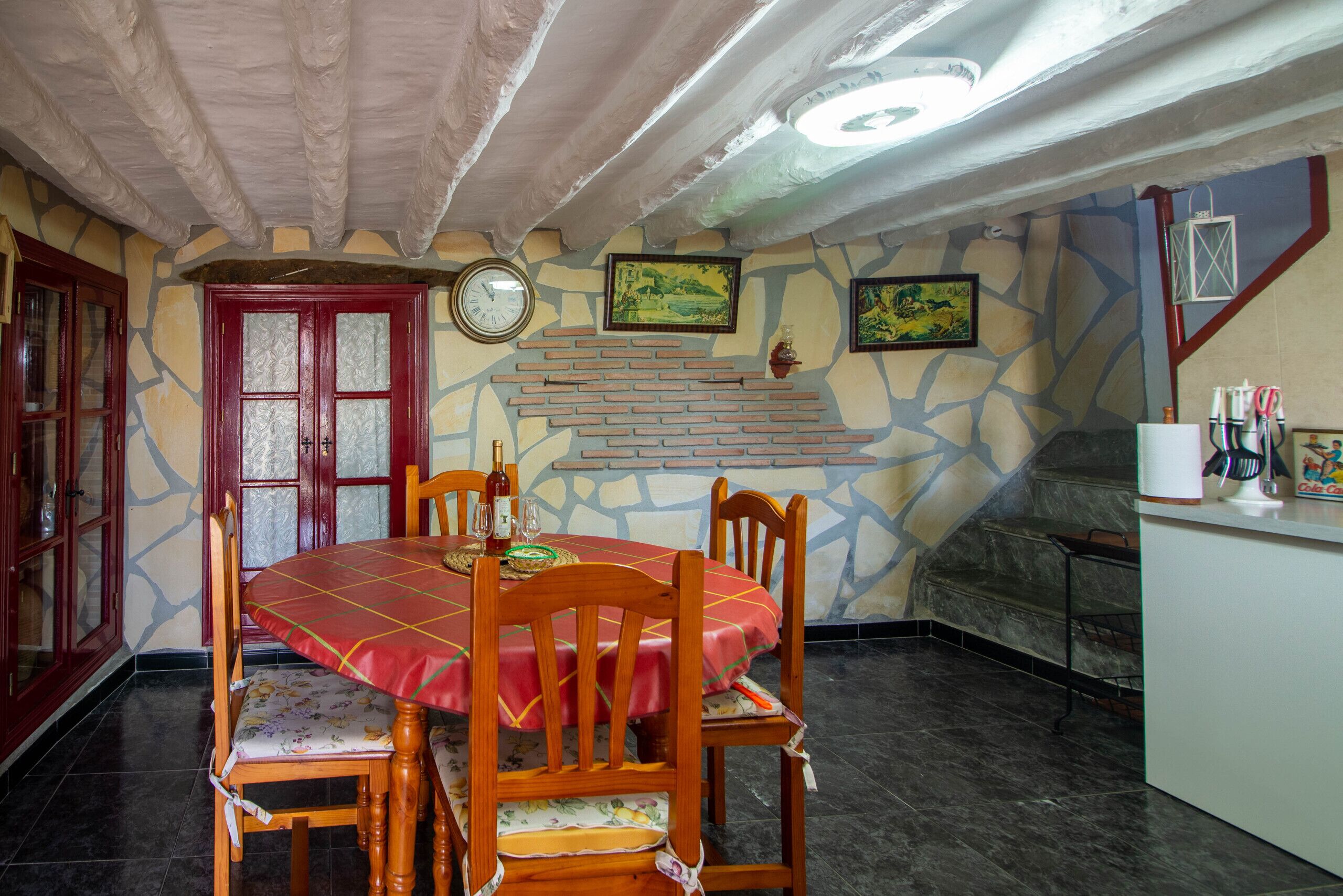 A dining area with a red tablecloth, wooden chairs, a clock, and a brick wall.