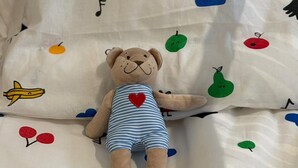 En blød bamse på en seng med et hvidt lagen med frugt- og sky mønstre.