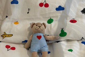 Ein kuscheliges Stofftier Bär auf einem Bett mit einer weißen Bettdecke, verziert mit Obst- und Wolkenmotiven.