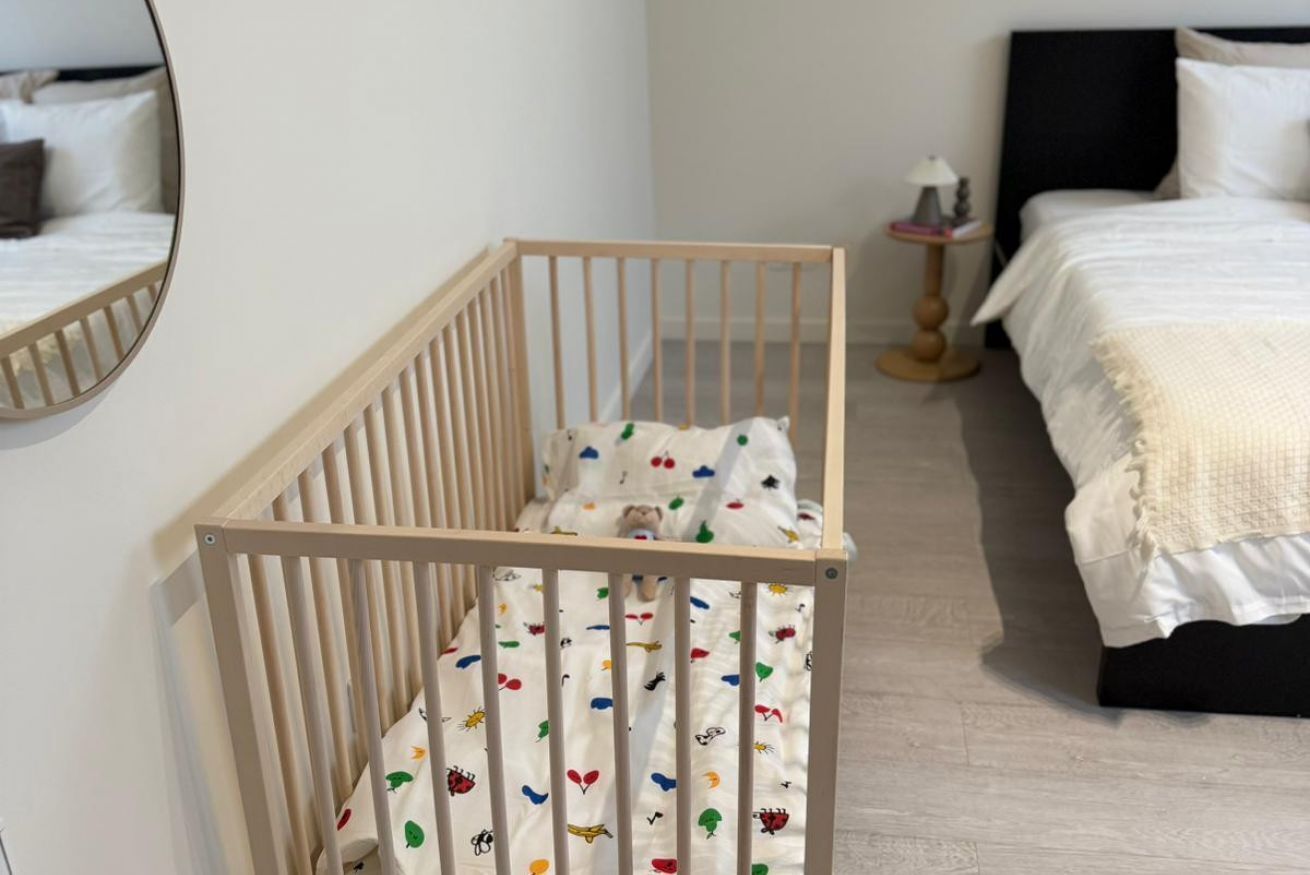 Um berço com colchão branco e estampas coloridas, uma mesa de cabeceira com abajur e uma cama com roupa de cama branca em um quarto com piso de madeira clara.