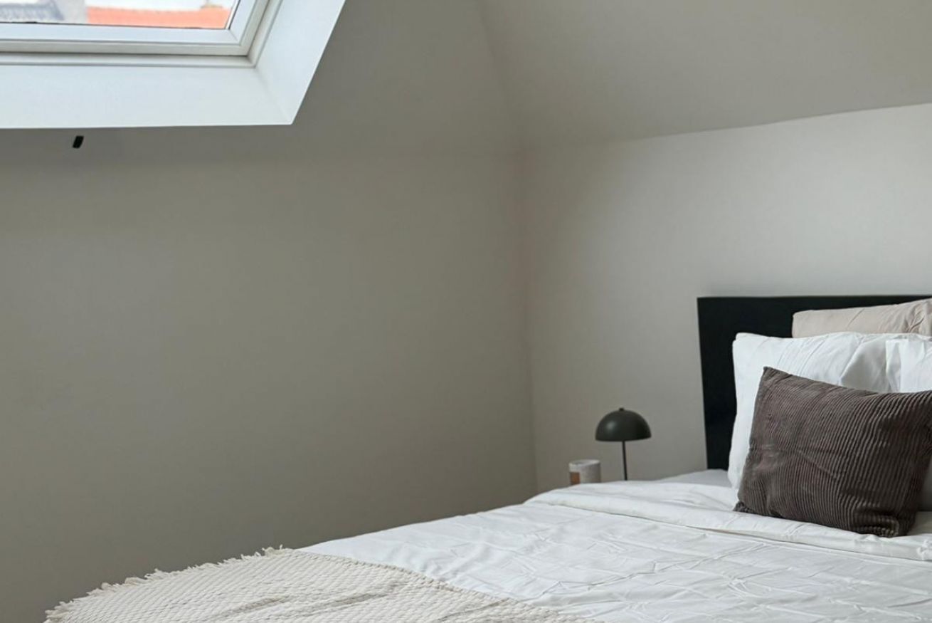 A bedroom with a skylight, a bed with white bedding and a brown pillow, a nightstand with a lamp, and a plain wall.