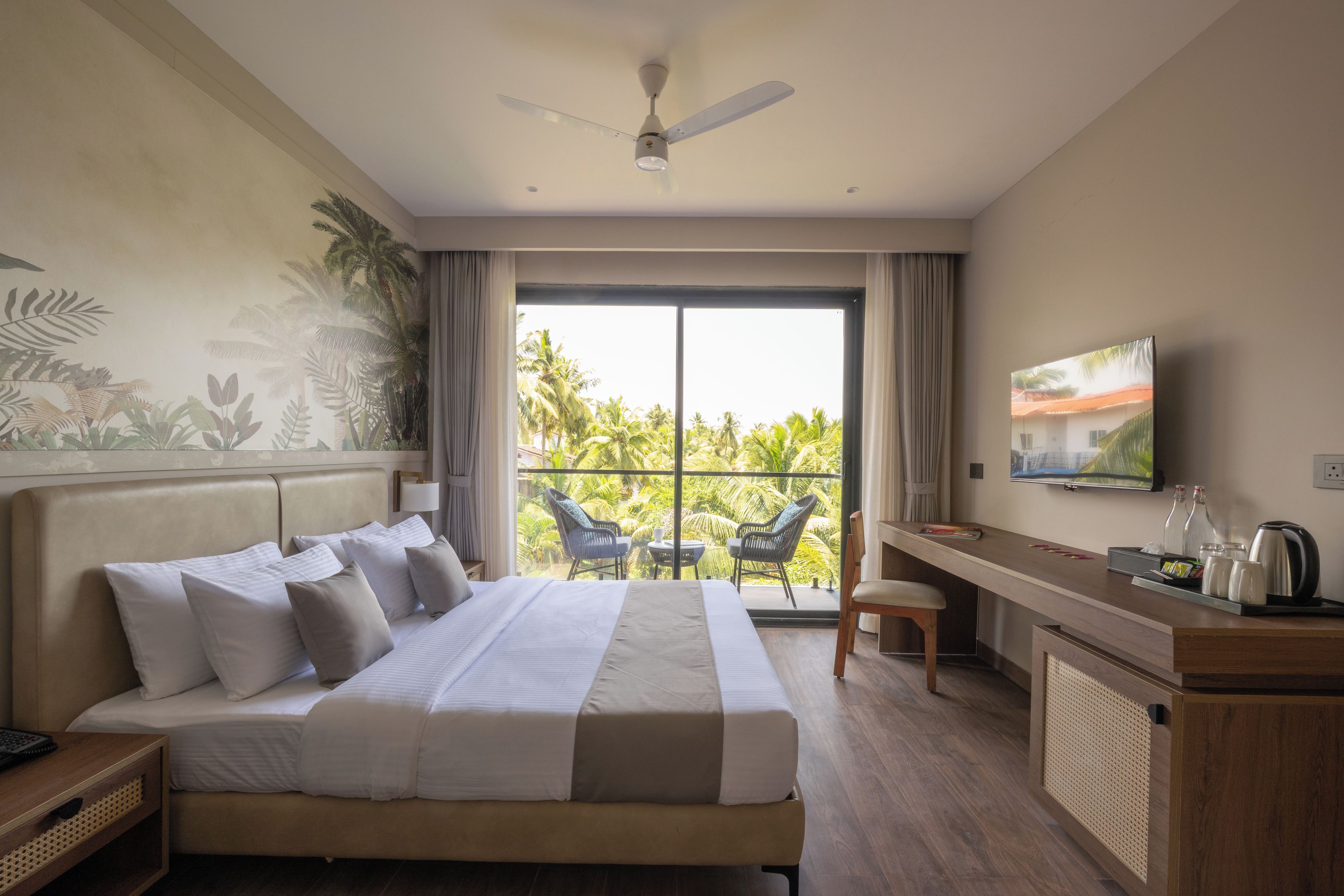 Una habitación de hotel con una cama grande, un escritorio, un televisor y un balcón con vista a palmeras.