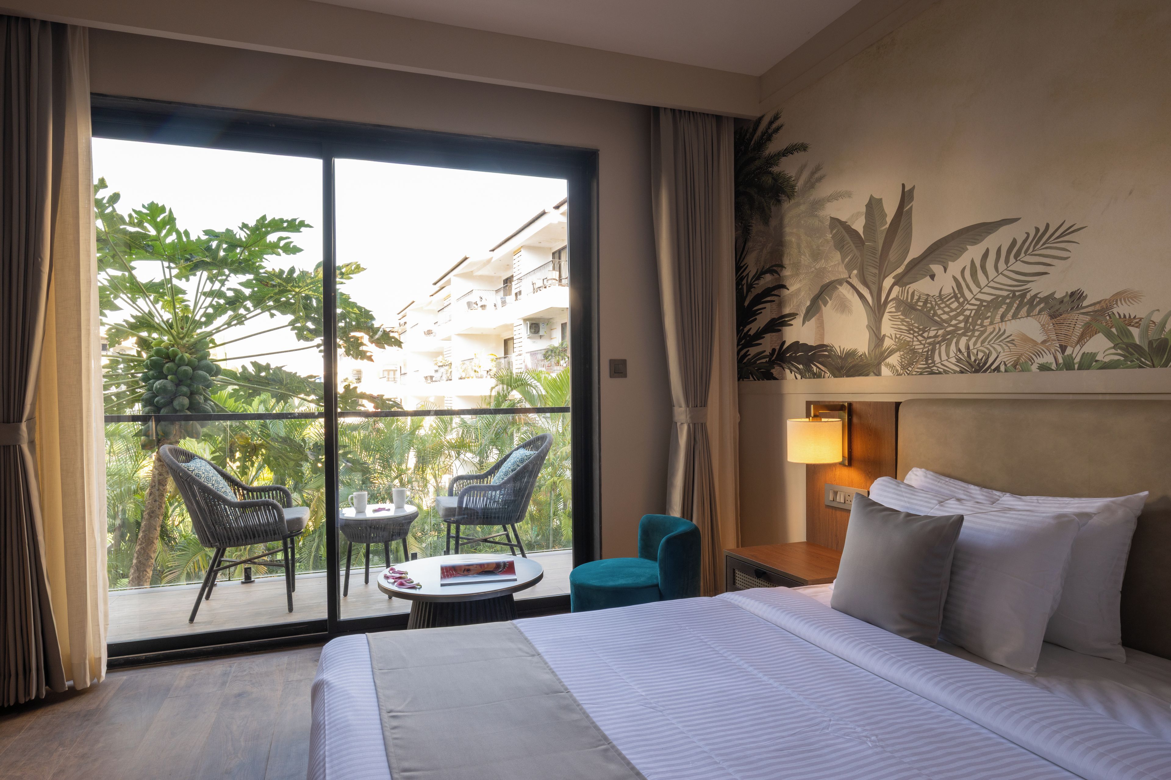 Habitación de hotel con cama, mesita de noche, lámpara, ventana con cortinas, balcón con sillas y vistas a zonas verdes y edificios.