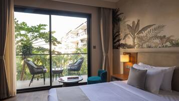 Habitación de hotel con cama, mesita de noche, lámpara, ventana con cortinas, balcón con sillas y vistas a zonas verdes y edificios.