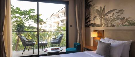 A hotel room with a bed, a nightstand, a lamp, a window with curtains, a balcony with chairs, and a view of greenery and buildings.