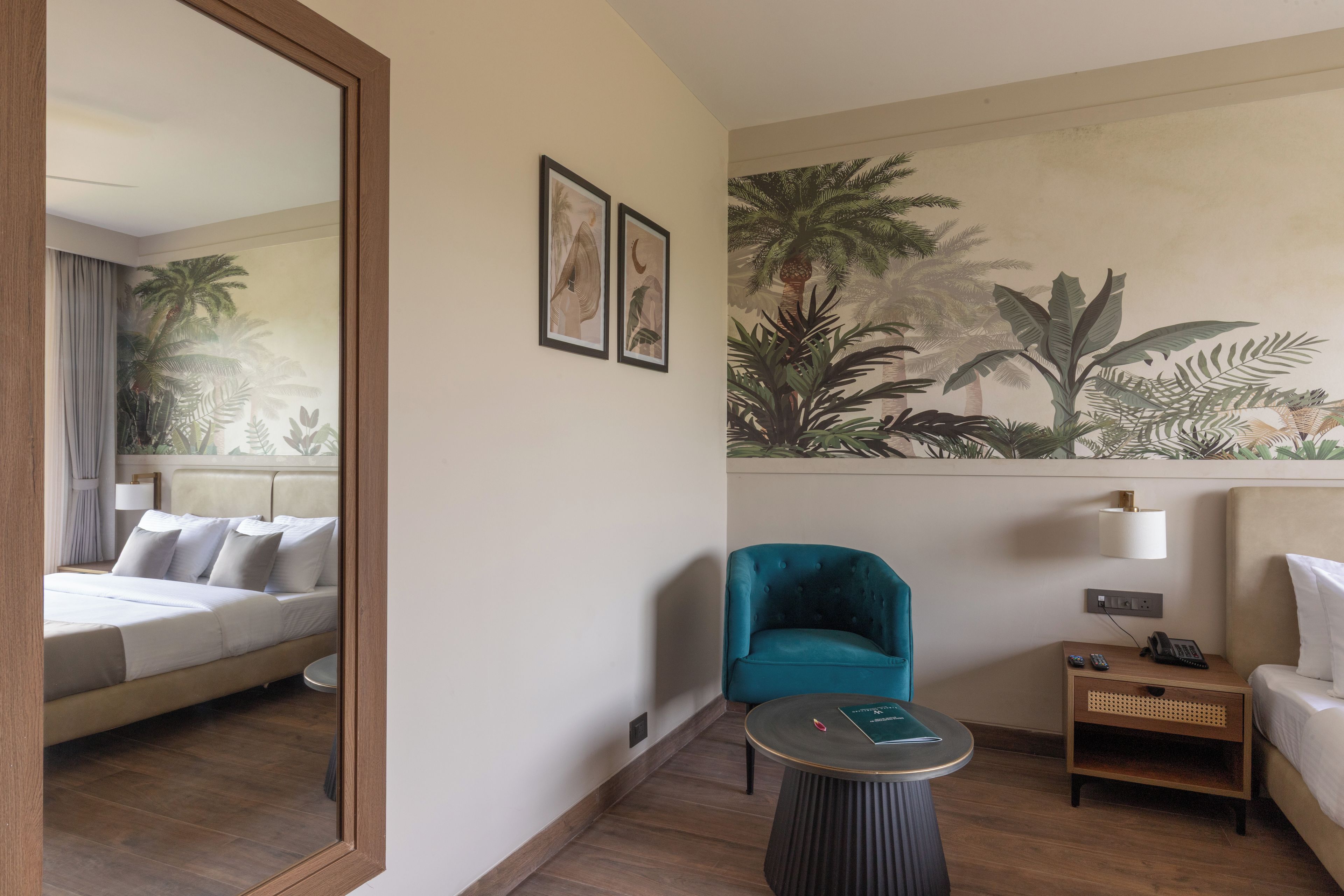 Quarto de hotel com cama, área de estar com poltrona e mesa pequena, mesa de cabeceira com luminária e um grande mural na parede com folhagem tropical.