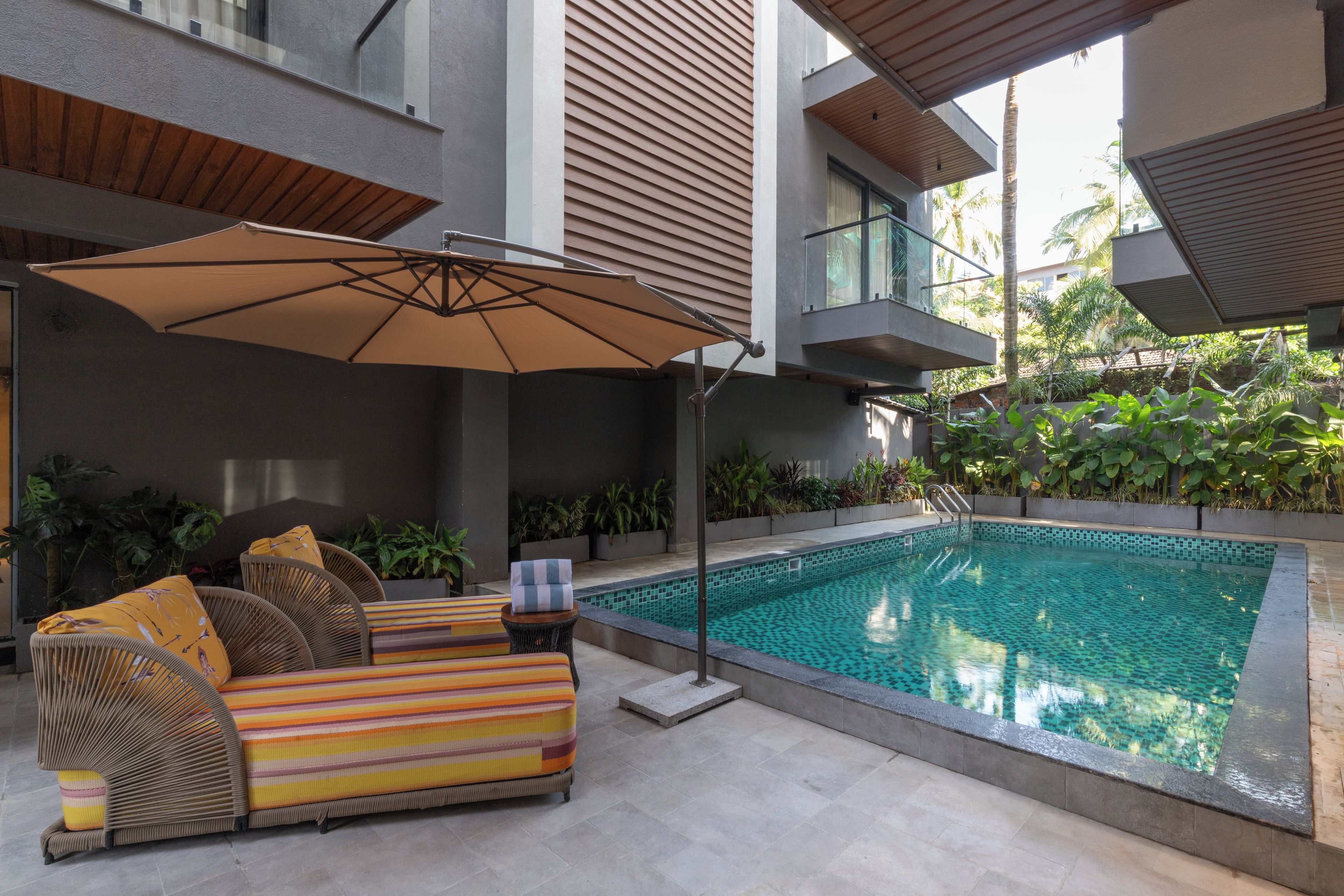 Un área de piscina moderna al aire libre con una silla de descanso y una sombrilla, rodeada de vegetación y un edificio con grandes ventanales.