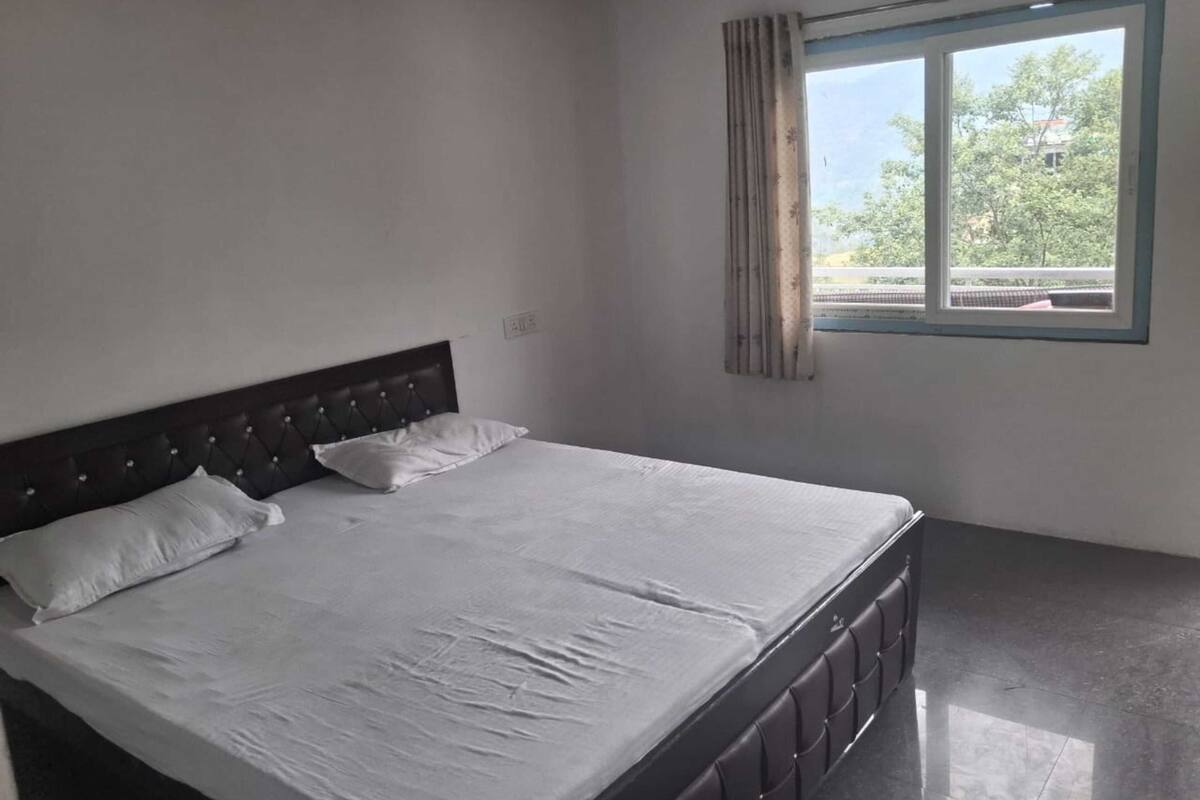 A single bed with a headboard, a window with curtains, and a view of trees and buildings outside.