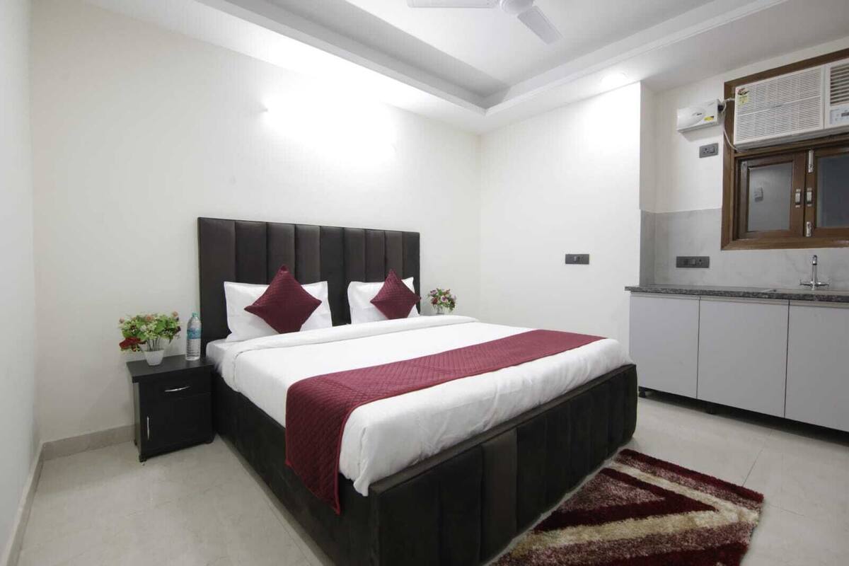 A bedroom with a bed, nightstand, dresser, and a kitchenette area.
