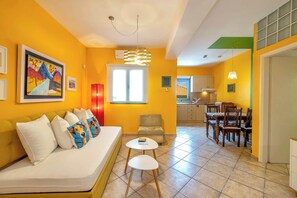 A living room with a sofa, a dining table, chairs, a small table with a book, a chair, a painting on the wall, and a window. - Alkmene apartment - Warm Getaway in Kefalonia (Agia Effimia)