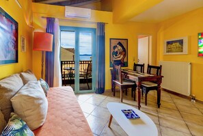 A living room with a sofa, a dining table with chairs, a painting, and a view through a glass door. - Phaedra apartment - Homely Vibes Gem in Kefalonia (Agia Effimia)
