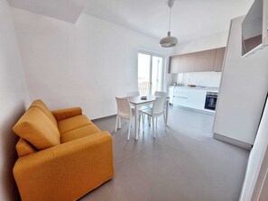 Ein modernes Wohnzimmer mit einer Couch, einem Esstisch mit Stühlen, einer Küchenzeile mit Schränken und Geräten sowie einem großen Fenster.