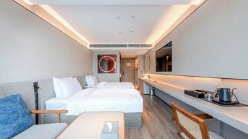A modern hotel room with two beds, a desk, a chair, a flat-screen TV, and a kitchenette.