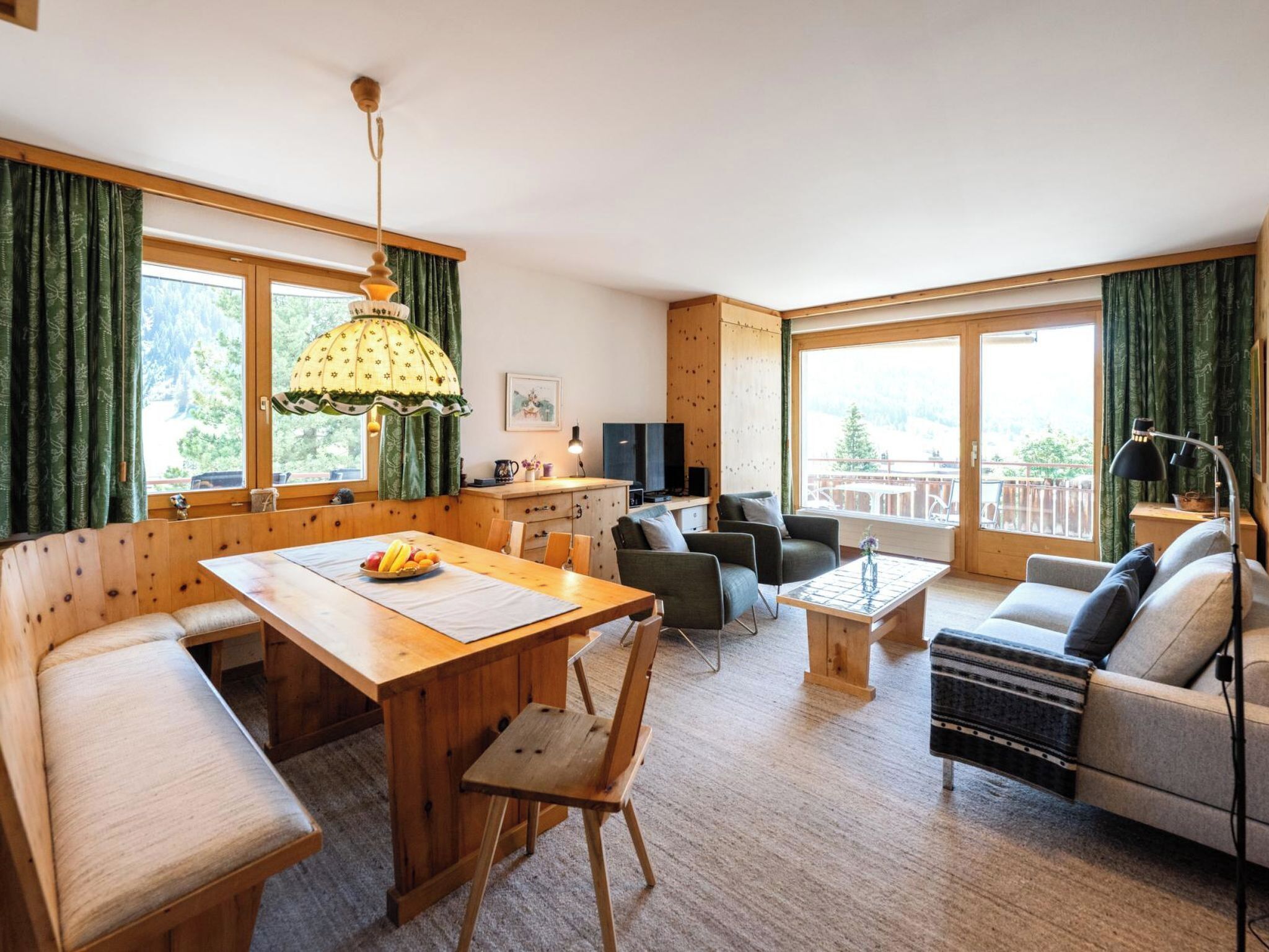 A cozy room with a wooden dining table, a sofa, a TV, and a balcony with a view.