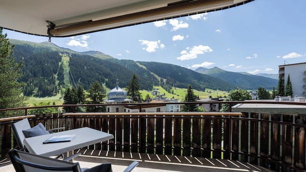 Un balcon avec une table et des chaises, donnant sur un paysage montagneux parsemé de bâtiments et de verdure.