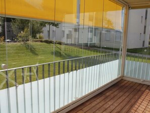 Ein Balkon mit Glasveranda, Holzboden und Blick auf eine Rasenfläche mit Gebäuden im Hintergrund.
