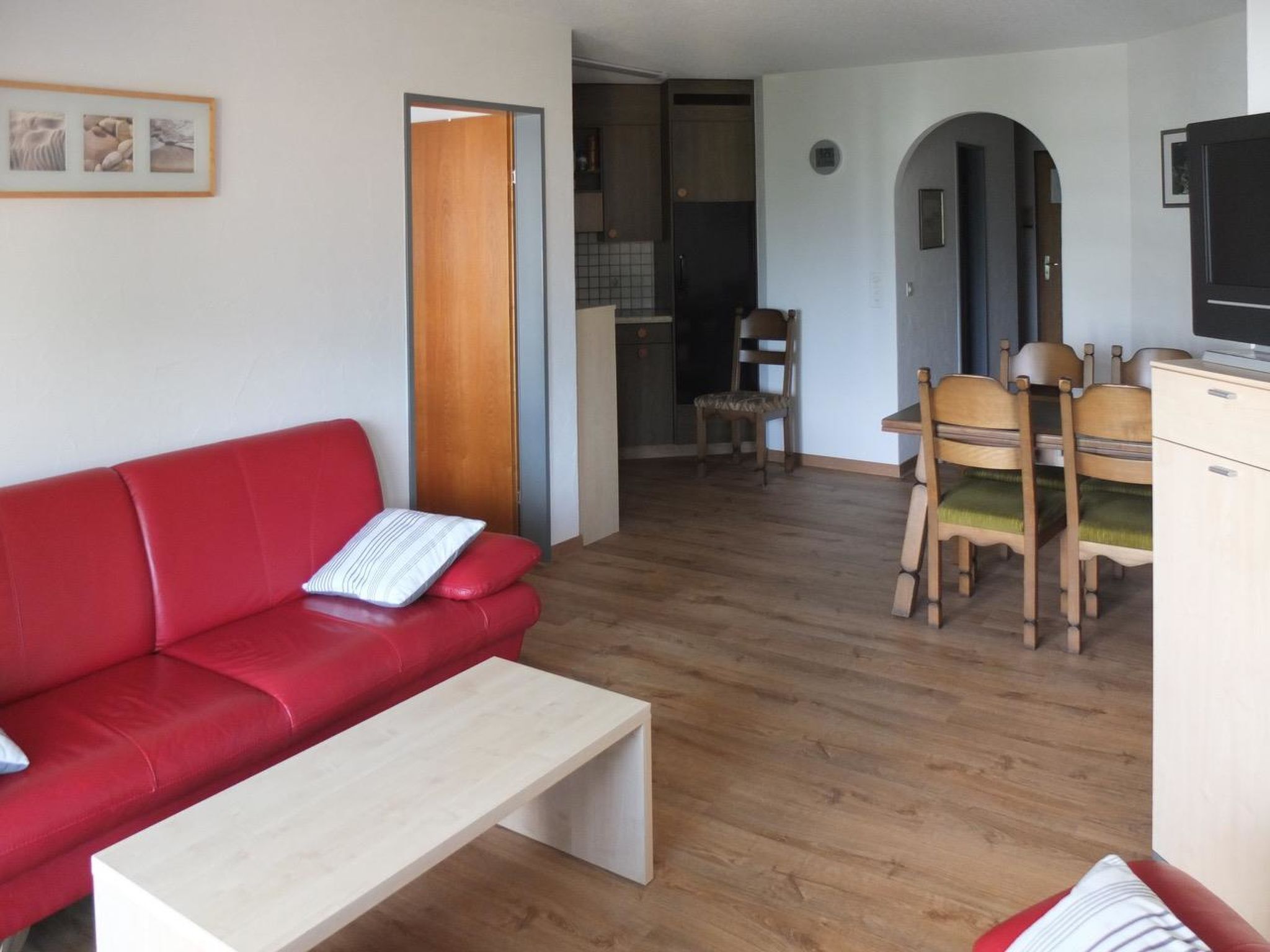 A living room with a red sofa, a wooden dining table with chairs, a kitchen area, and a TV on a cabinet.