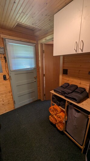 A small room with a wooden ceiling, a door with a window, a bed with a headboard, and a shelf with towels.