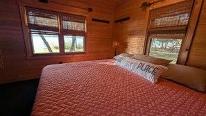 A wooden cabin bedroom with a bed, two windows, a lamp, and a sign on the wall.