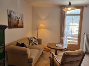 Ein Wohnzimmer mit einer Couch, einem runden Tisch, einer Lampe, einem Bild von einem Reh und einem Fenster mit Vorhängen.
