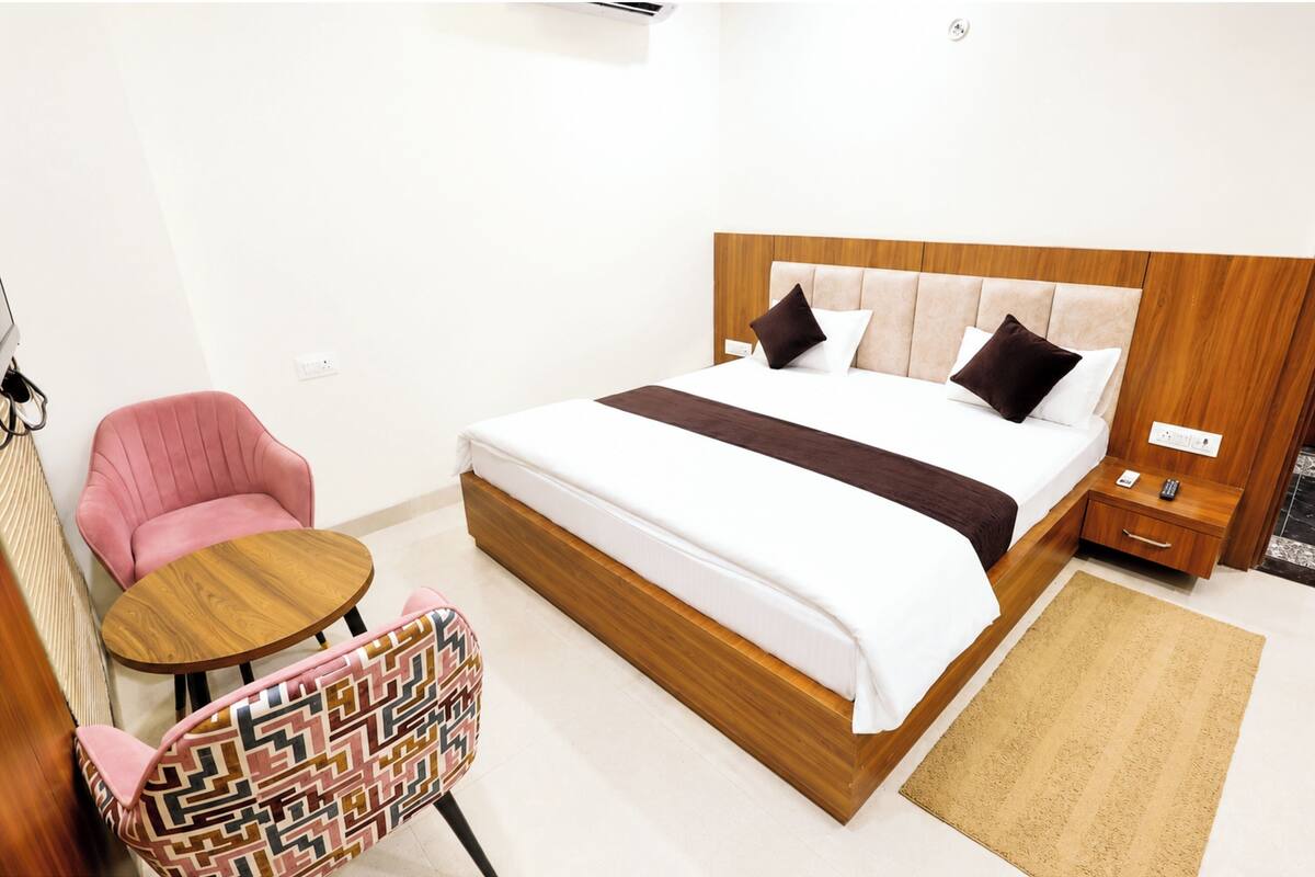 A hotel room with a bed, a small round table, a pink chair, and a patterned armchair.