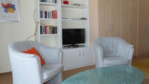 Una sala de estar con dos sillones blancos, una mesa de centro de vidrio, una estantería con libros y un televisor, y un armario de madera.