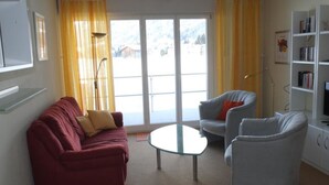 Una sala de estar con un sofá rojo, un sillón blanco, una mesa de centro de vidrio y una estantería.