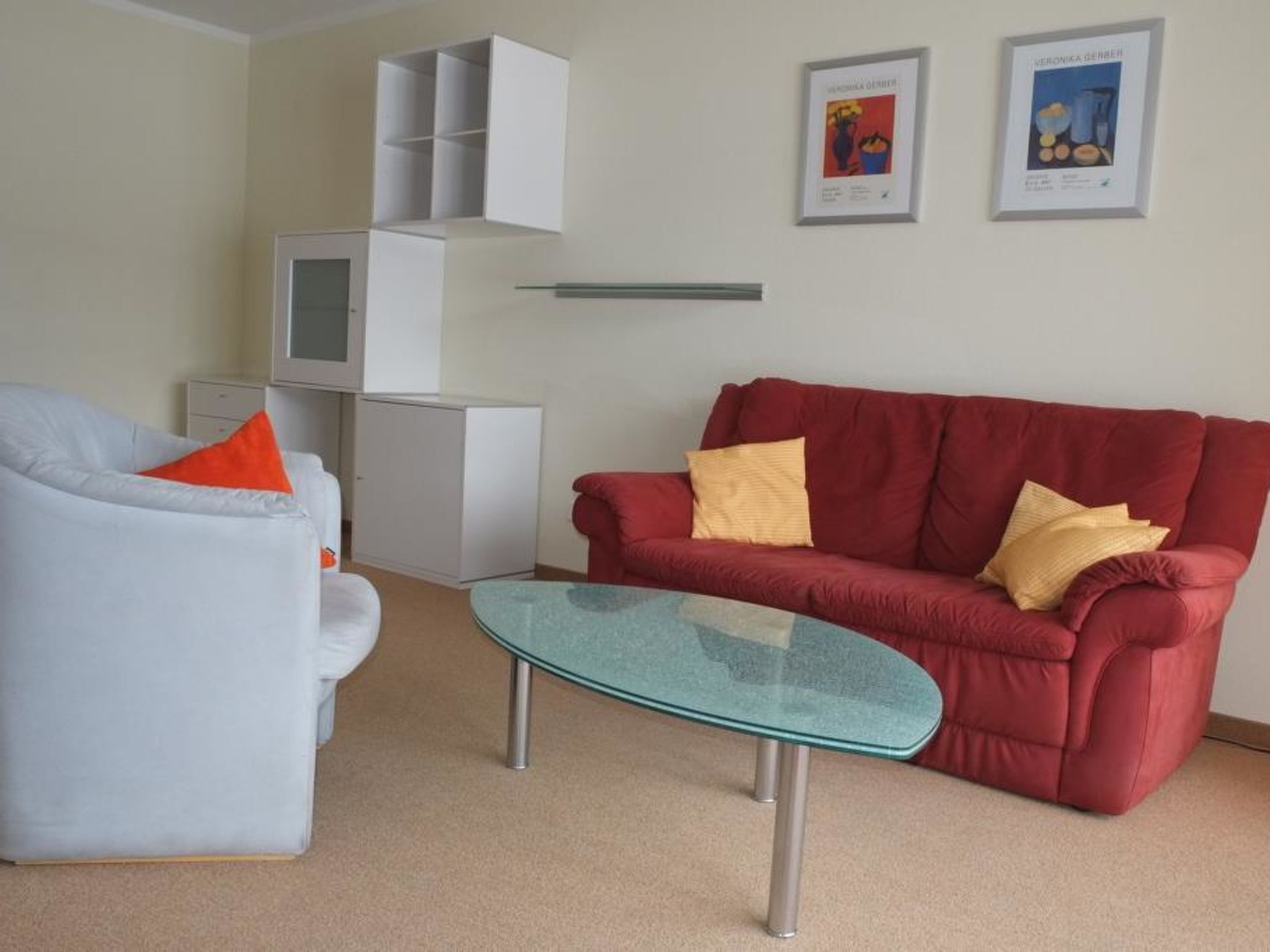 Een woonkamer met een rode bank, een witte fauteuil, een glazen salontafel, een wit kastje en twee ingelijste kunstwerken aan de muur.