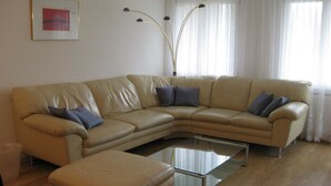 Un canapé d’angle en cuir beige, accompagné d’une table basse en verre, d’un petit pouf et d’un grand lampadaire.