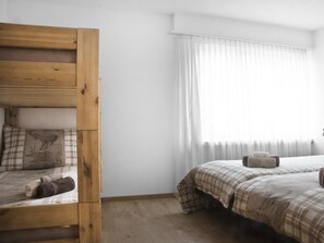 Ein Schlafzimmer mit Etagenbett, zwei Betten, einem Fenster mit durchsichtigen Vorhängen und einer Heizung.