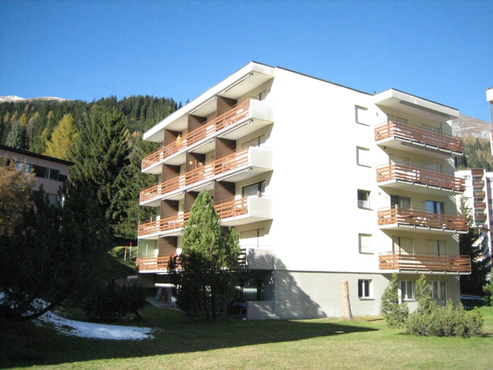 A multi-story building with balconies, surrounded by trees and a mountain.