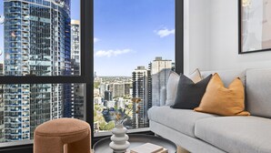 A modern living room with a sofa, cushions, a round table with a vase and books, and a large window with a city view. - Bright Apt Southbank Near Crown Casino ,pool &gym (Melbourne)
