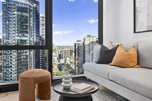 A modern living room with a sofa, cushions, a round table with a vase and books, and a large window with a city view. - Bright Apt Southbank Near Crown Casino ,pool &gym (Melbourne)