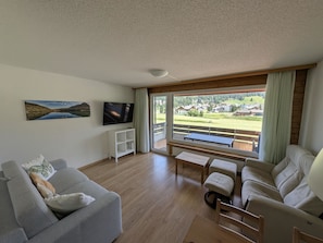 Ein Wohnzimmer mit Fernseher, Sofa und Blick auf eine Wohngegend durch große Fenster.