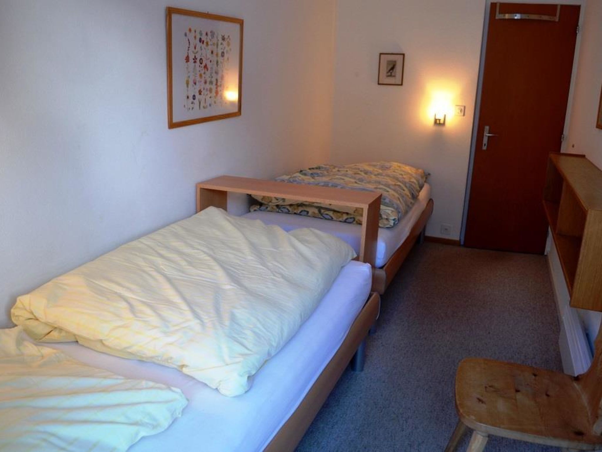 A small, single-room accommodation with two beds, a wooden desk, a chair, and a door.