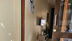 A hallway with a tiled floor, a wooden door, a small table with a vase, and a wall-mounted TV. - Stylish Studio Apartment In Fourways (Sandton)