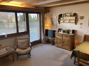 A cozy room with a wooden chair, a small table with books, a TV, a cabinet, and a window with lace curtains. - Albeina Hus Appartement/Fewo, Dusche oder Bad, WC 2-Zimmerwohnung by Interhome (Klosters)
