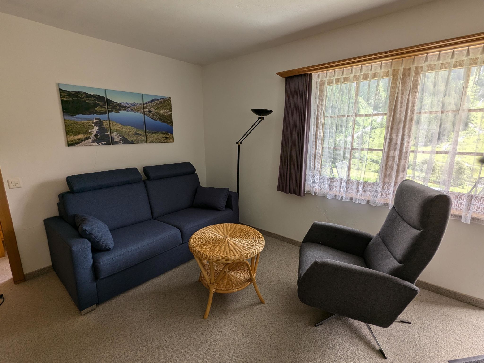 Un salon avec un canapé bleu foncé, une table d’appoint en osier, un fauteuil gris, une lampe sur pied et une fenêtre ornée de rideaux légers.