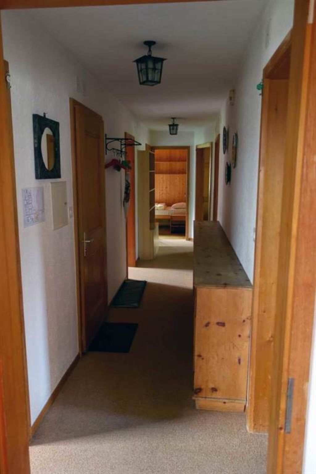 Un couloir étroit avec des portes en bois, un lit au fond et un luminaire au plafond.