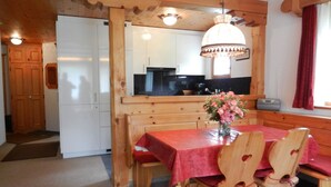 A wooden dining area with a table and chairs, a hanging light fixture, and a kitchen in the background.