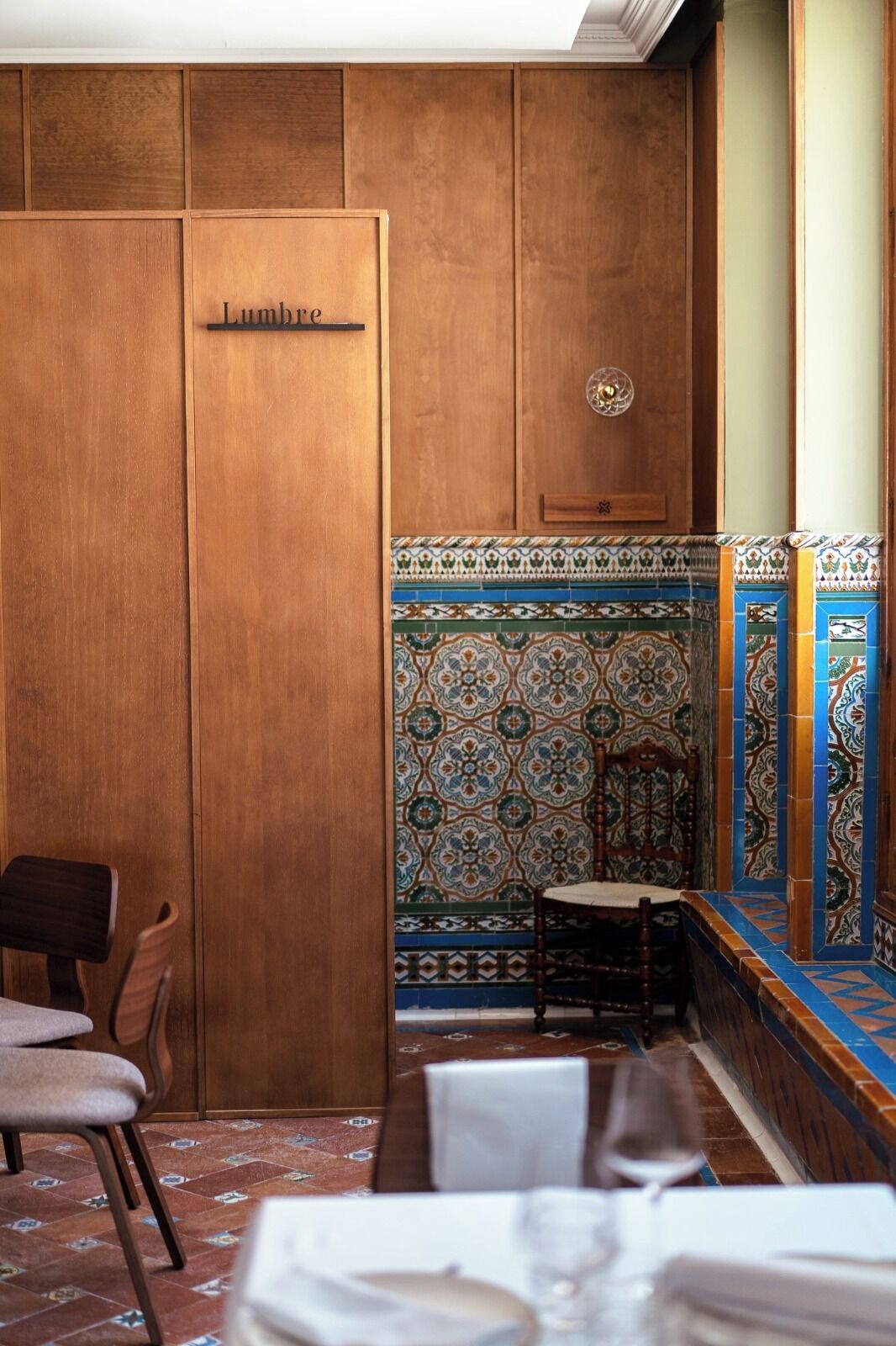 Une pièce avec une porte en bois portant l’inscription « Lumme », un mur recouvert de carreaux décoratifs, une chaise en bois et une table recouverte d’une nappe blanche.