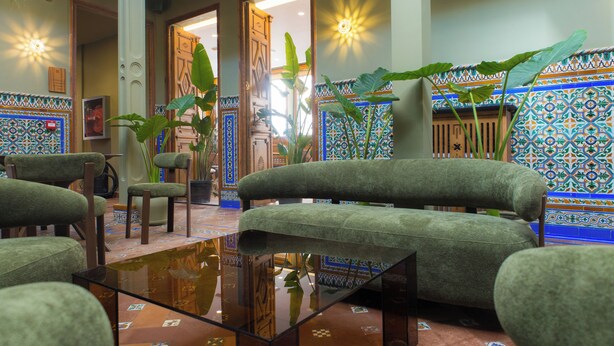 A lobby with a green sofa, chairs, a glass coffee table, and a tiled wall with a geometric pattern.